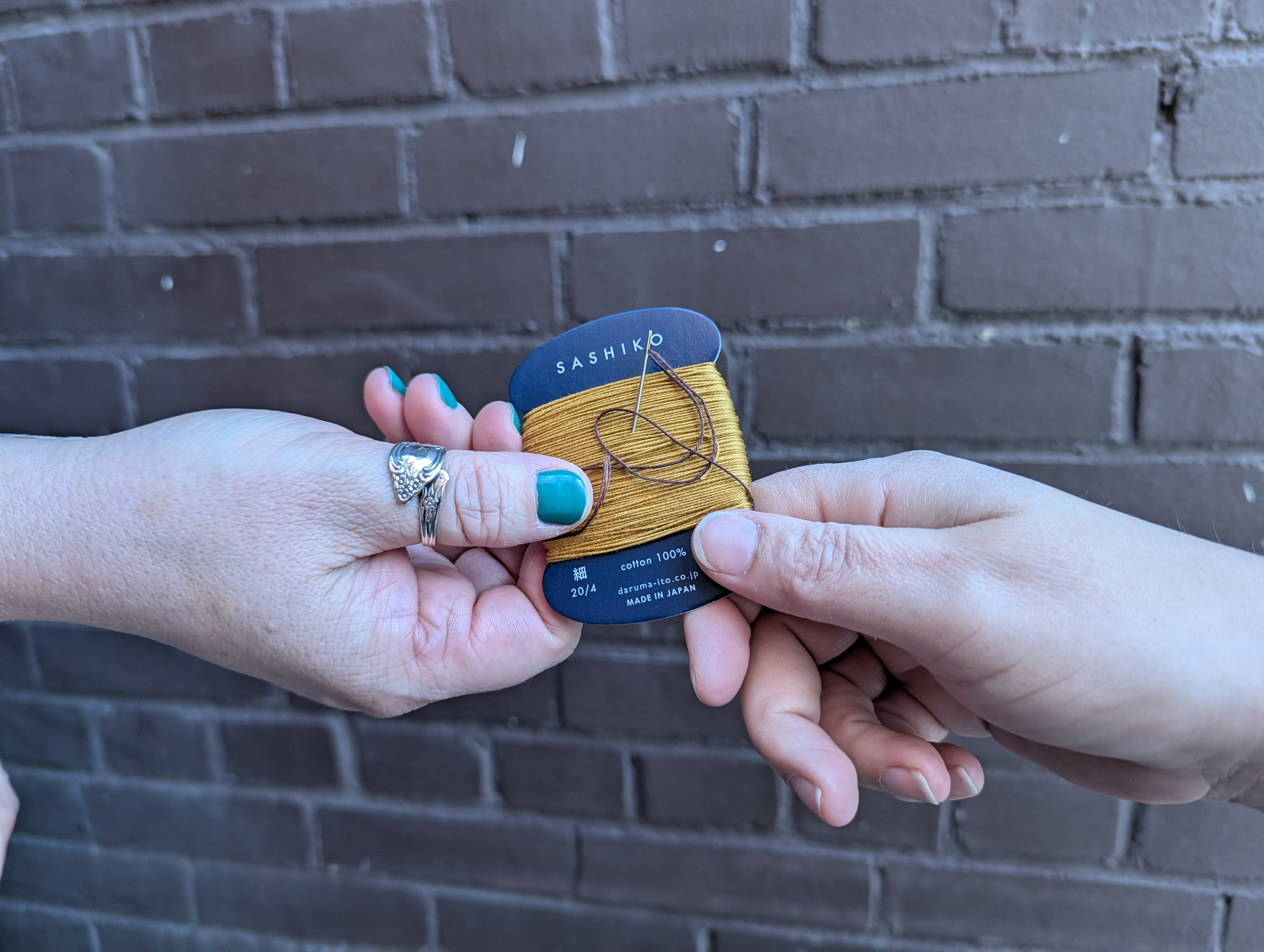 One person's hand giving a spool of sashiko thread to another outstretched hand